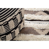 A black and white shaggy pouffe featuring a geometric pattern, placed on a textured, light-colored rug with subtle designs. The pouffe has a round shape and a soft, fluffy texture, contrasting with the smooth surface of the rug underneath