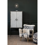 A black and white shaggy pouffe placed in a stylish living room setting, featuring a dark green wall, a light-colored wooden floor, and a patterned armchair nearby. A tall cabinet with a circular handle is visible in the background, along with a decorative light fixture above
