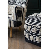 A black and white shaggy pouffe with a textured design, featuring tassels along the bottom edge, positioned next to a patterned armchair with wooden legs. The background includes a dark green wall and a wooden floor