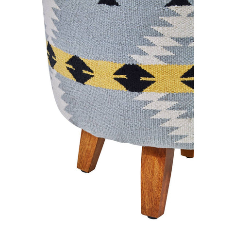 Round footstool featuring a colorful Kilim-style fabric with geometric patterns in blue, black, and yellow. The footstool is supported by four sturdy mango wood legs, showcasing a natural wood finish