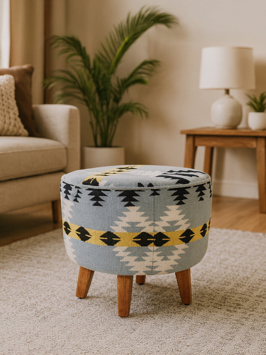 Round footstool featuring a colorful Kilim-style fabric with geometric patterns in blue, yellow, and black, supported by four sturdy mango wood legs. The footstool is placed on a light-colored rug in a cozy living room setting, with a plant and lamp visible in the background