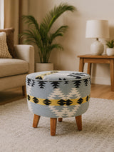 Round footstool featuring a colorful Kilim-style fabric with geometric patterns in blue, yellow, and black, supported by four sturdy mango wood legs. The footstool is placed on a light-colored rug in a cozy living room setting, with a plant and lamp visible in the background