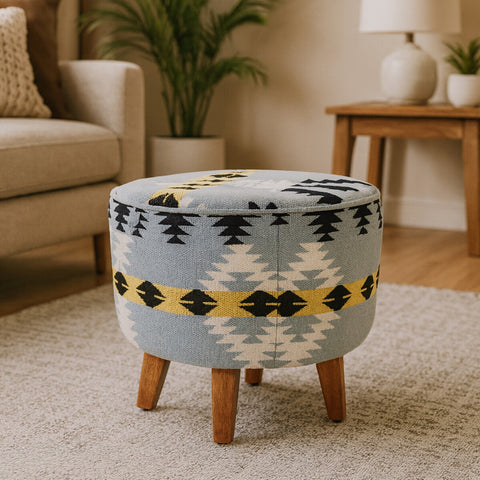 Round footstool featuring a colorful Kilim-style fabric with geometric patterns in blue, yellow, and black, supported by four tapered mango wood legs. The footstool is placed on a light-colored rug in a cozy living room setting with a sofa and a plant in the background