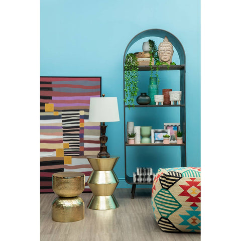 A colorful kilim-style pouffe featuring geometric patterns in vibrant hues, positioned next to a modern bookshelf filled with books and decorative items, alongside a stylish lamp and metallic side tables, all set against a bright blue wall