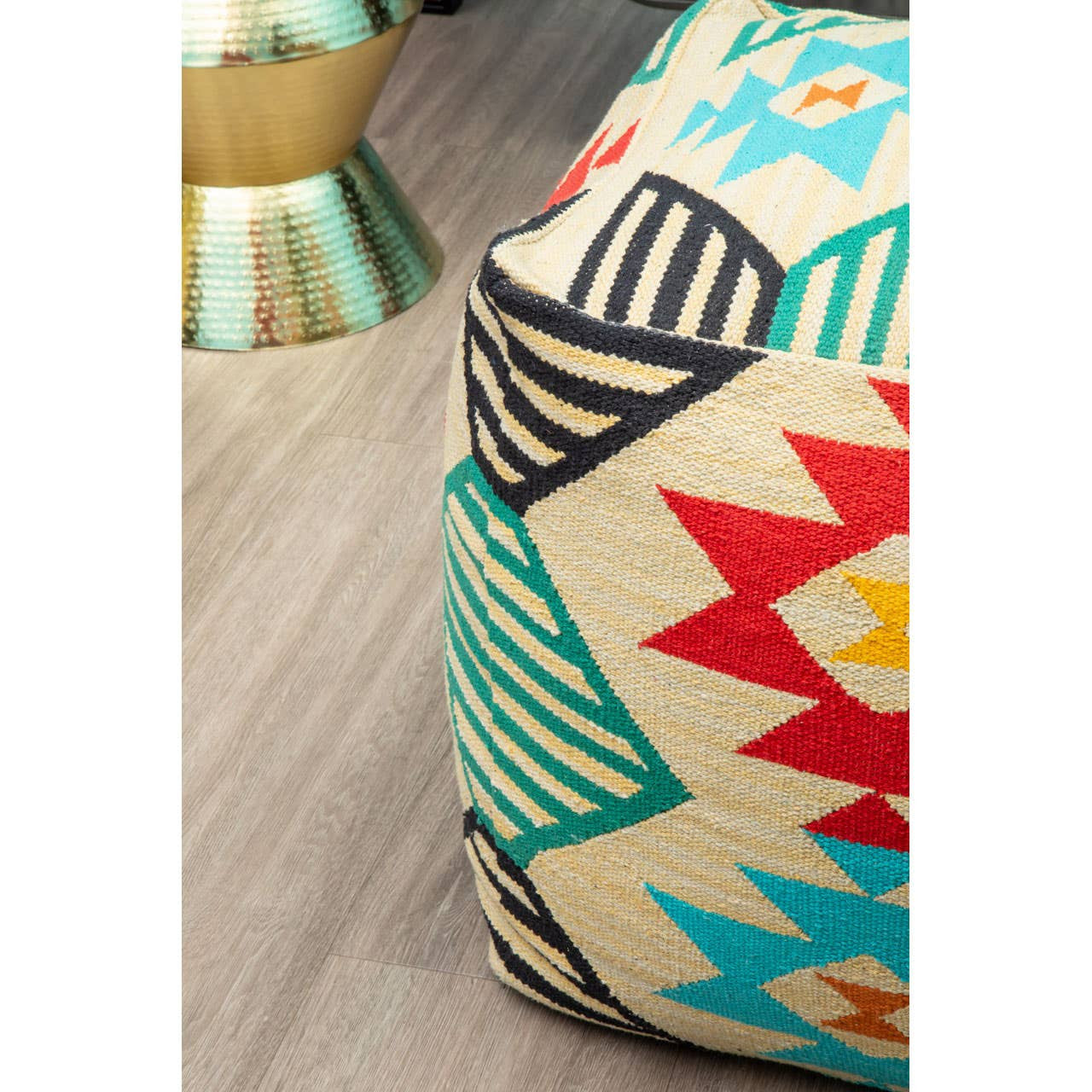 A colorful kilim style pouffe featuring a geometric pattern with red, blue, yellow, and black accents. The pouffe is placed on a light wooden floor, with a metallic decorative item partially visible in the background