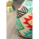 A colorful kilim style pouffe featuring a geometric pattern with red, blue, yellow, and black accents. The pouffe is placed on a light wooden floor, with a metallic decorative item partially visible in the background