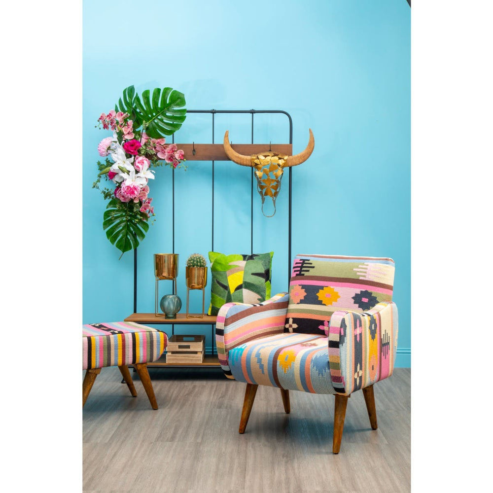 Multicolour footstool featuring a vibrant, patterned upholstery with various geometric and floral designs, complemented by wooden legs. The footstool is positioned in a bright room with a blue wall, alongside a matching chair and decorative elements like a plant and wall decor