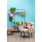 Multicolour footstool featuring a vibrant, patterned upholstery with various geometric designs, complemented by wooden legs. The footstool is placed in a bright room with a blue wall, alongside a matching chair and decorative elements like plants and a wall-mounted rack