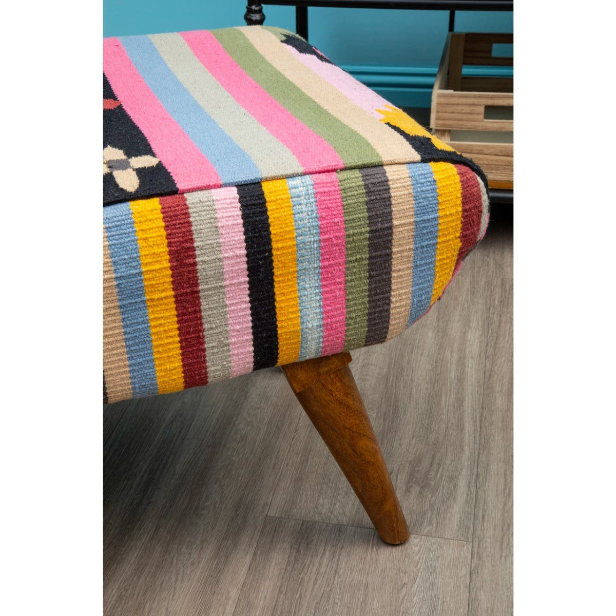 Multicoloured footstool featuring a striped fabric design in various vibrant colors, complemented by wooden legs. The footstool has a rounded top and a sturdy, angled leg structure, set against a light wooden floor