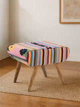 Multicolour footstool with a striped fabric design featuring vibrant hues of pink, blue, yellow, and green, supported by four wooden legs. The footstool is placed on a light-colored rug in a bright room with a neutral wall and a framed artwork in the background