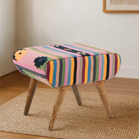 A multicoloured footstool featuring a vibrant striped pattern in shades of pink, orange, green, and blue. The footstool has a cushioned top with rounded edges and is supported by four wooden legs. It is placed on a light-colored woven rug in a cozy interior setting