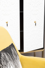 A stylish black and white two-door cabinet featuring sleek white doors with gold handles, positioned next to a yellow accent chair with a decorative black and white pillow. The cabinet has a modern design and is set against a neutral background