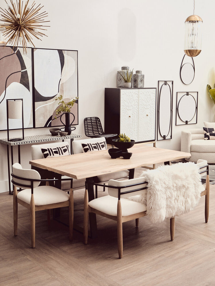 A stylish dining area featuring a light wood table surrounded by four upholstered chairs with white cushions. In the background, a black and white two-door cabinet stands against a neutral wall, complemented by decorative wall art and a round mirror. The space is bright and modern, with a minimalist aesthetic