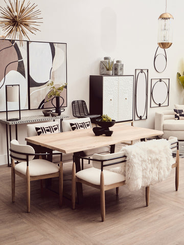 A stylish dining area featuring a light wood table surrounded by four upholstered chairs with white cushions. In the background, a black and white two-door cabinet stands against a neutral wall, complemented by decorative wall art and a round mirror. The space is bright and modern, with a minimalist aesthetic