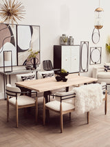 A stylish dining area featuring a light wood table surrounded by four upholstered chairs with white cushions. In the background, a black and white two-door cabinet stands against a neutral wall, complemented by decorative wall art and a round mirror. The space is bright and modern, with a minimalist aesthetic
