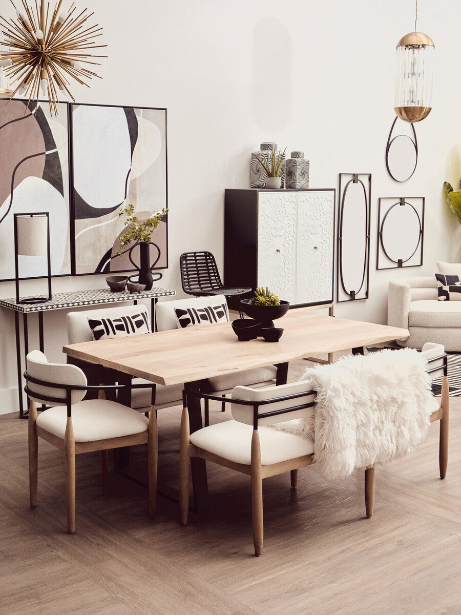 A stylish dining area featuring a light wood table surrounded by four upholstered chairs with white cushions. In the background, a black and white two-door cabinet stands against a neutral wall, complemented by decorative wall art and a round mirror. The space is bright and modern, with a minimalist aesthetic