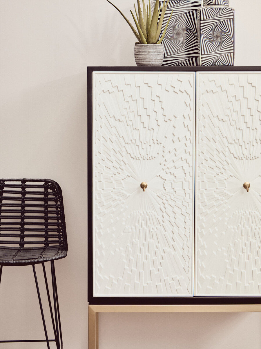 Dumas Black and White Two Door Cabinet featuring a textured white front with two round gold knobs, set against a black frame and supported by light wooden legs, alongside a modern black metal stool and a small potted plant on top
