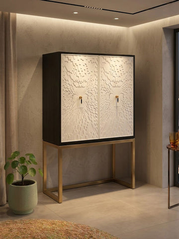 A stylish two-door cabinet featuring a black frame and white doors with intricate designs, set on a sleek gold metal base. The cabinet is positioned against a softly lit wall, with a potted plant nearby, creating a modern and elegant home decor setting