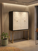 A stylish two-door cabinet featuring a black frame and white doors with intricate designs, set on a sleek gold metal base. The cabinet is positioned against a softly lit wall, with a potted plant nearby, creating a modern and elegant home decor setting