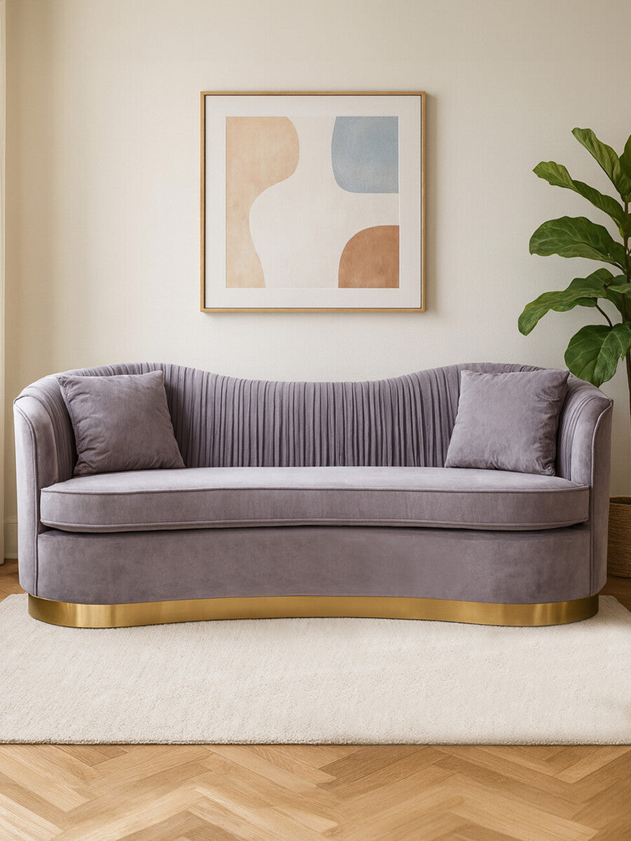 Three-seat pleated velvet sofa in grey with a curved backrest and two matching cushions, set against a light-colored wall. A framed abstract artwork hangs above the sofa, and a green plant is positioned to the right. The sofa features a gold base and is placed on a light-colored rug over a wooden floor