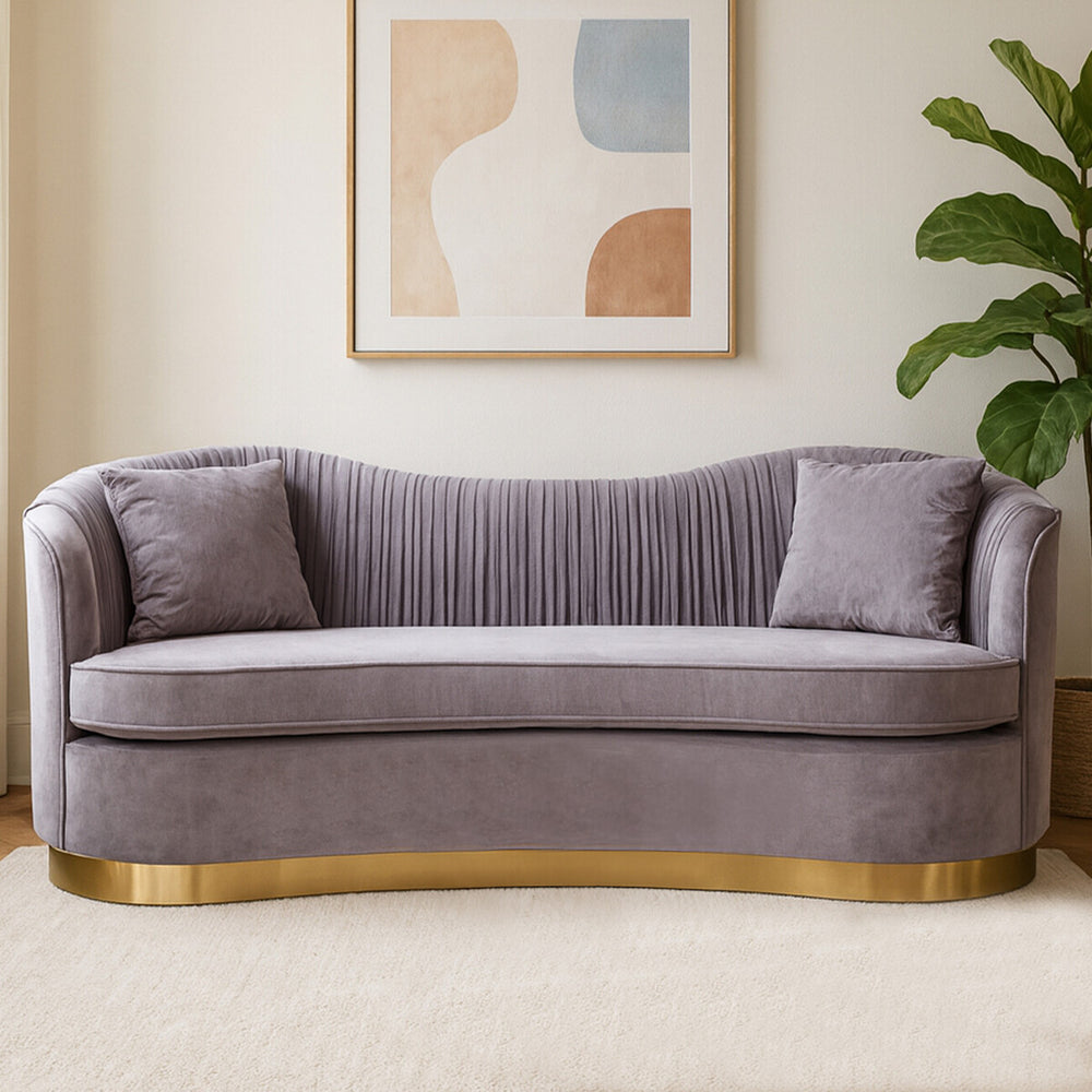 A three-seat sofa upholstered in soft grey pleated velvet, featuring two matching cushions and a sleek gold base. The sofa has a curved backrest and a modern design, set against a light-colored wall with abstract art and a green plant in the background