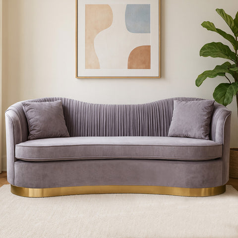 A three-seat sofa upholstered in soft grey pleated velvet, featuring two matching cushions and a sleek gold base. The sofa has a curved backrest and a modern design, set against a light-colored wall with abstract art and a green plant in the background