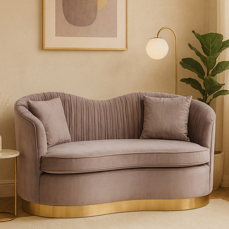 A grey pleated velvet sofa with a curved backrest and two matching cushions, featuring a gold base, set against a light-colored wall with a framed abstract artwork and a green plant in the background
