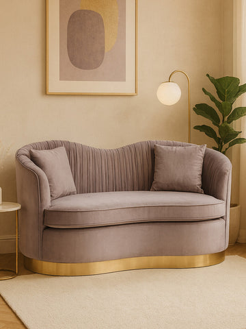 A grey pleated velvet sofa with a curved backrest and two matching cushions, featuring a gold base, set against a light-colored wall with a framed abstract artwork and a green plant in the background
