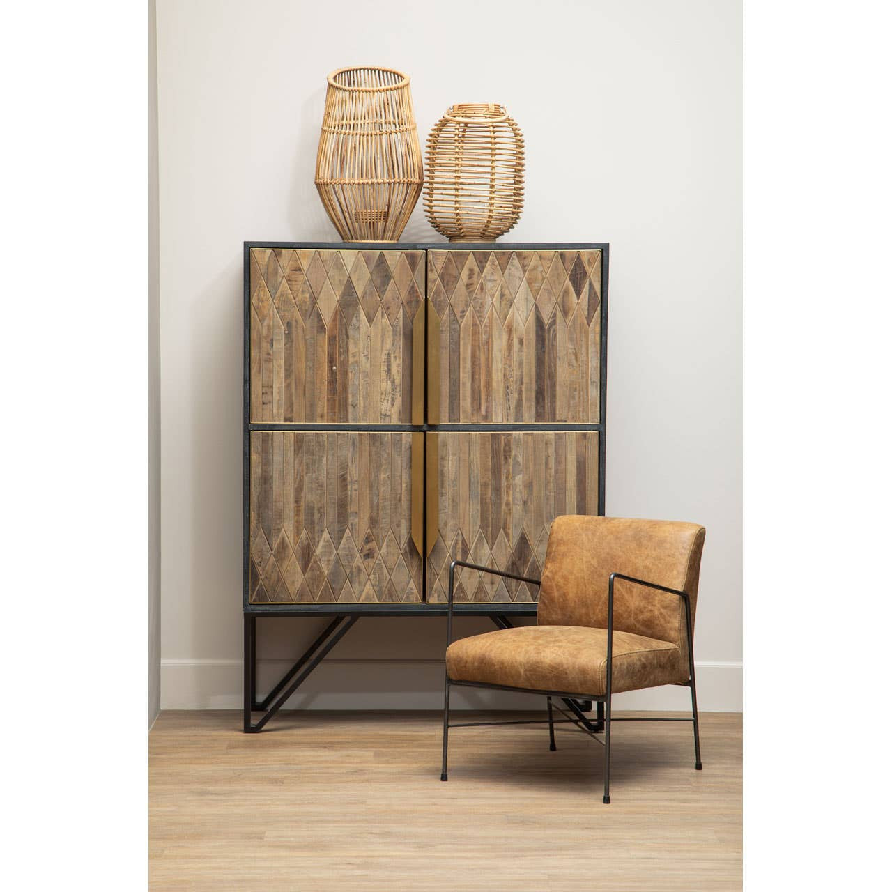 Textured wooden cabinet with a natural finish, featuring two large doors with a woven design, standing on a black metal frame. Above the cabinet are two decorative woven baskets. Next to the cabinet is a modern, tan upholstered chair with slender metal legs, set against a light-colored wall and wooden floor