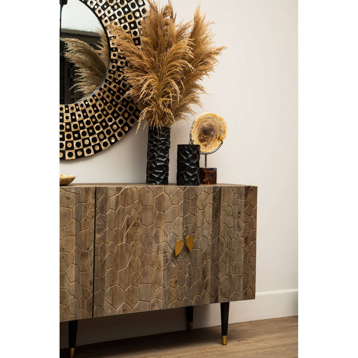 Tall cabinet made of recycled elm wood with a natural finish, featuring two doors and a sleek design. The cabinet is complemented by decorative items on top, including a large round mirror, a black vase with dried pampas grass, and a vintage-style speaker. The setting has a light-colored wall and wooden flooring, creating a warm and inviting atmosphere