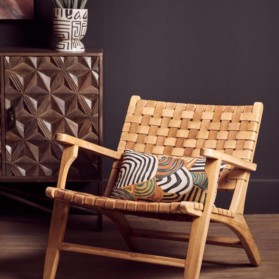 Wooden lounge chair with woven seat, patterned cushion, beside carved cabinet and plant pot