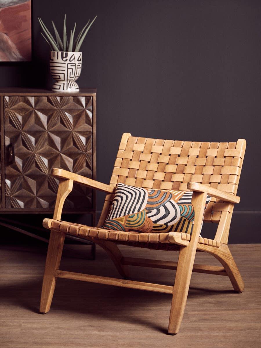 Wooden lounge chair with woven seat, patterned cushion, beside carved cabinet and plant pot