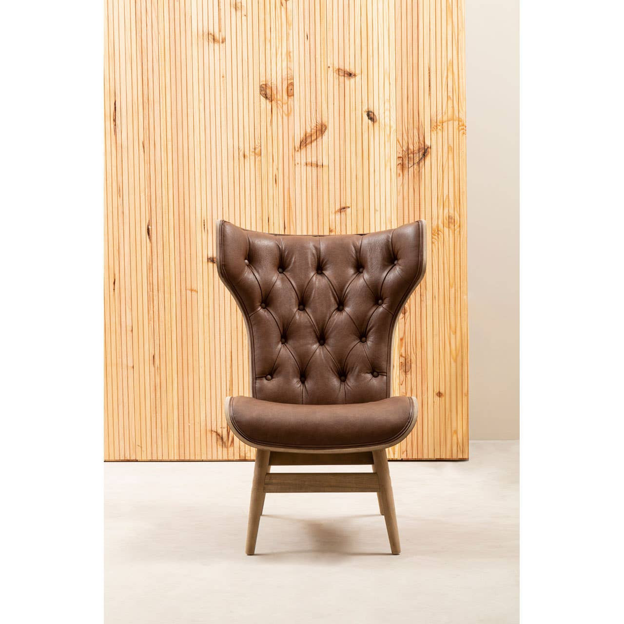 Brown leather effect chair featuring a winged back design with tufted detailing. The chair is supported by light wooden legs, providing a modern yet classic aesthetic.