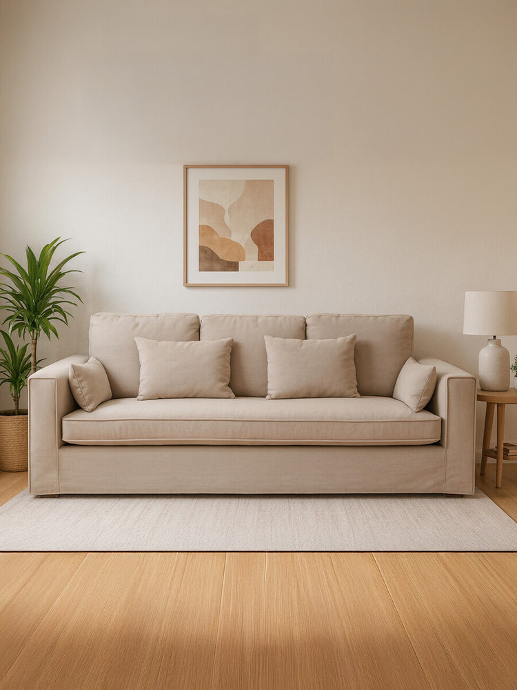 Three-seat sofa upholstered in light grey fabric, featuring plush cushions and wide armrests. The sofa is positioned against a neutral wall with a piece of abstract art above it. A potted plant is visible to the left, and a soft area rug is placed on a wooden floor. A lamp is situated on the right side of the sofa