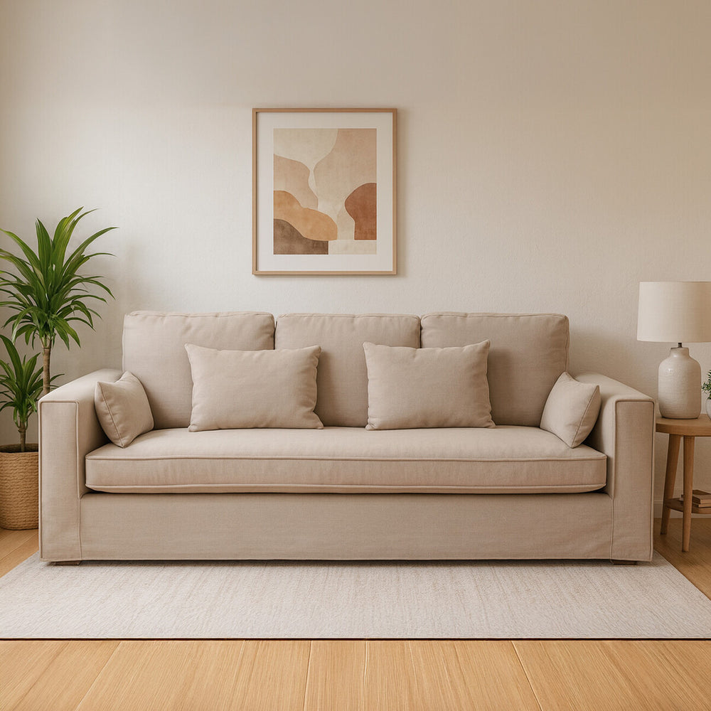 Three-seat grey fabric sofa with plush cushions, set against a light-colored wall. A framed artwork hangs above the sofa, and a potted plant is positioned to the left. A soft area rug is placed underneath the sofa, enhancing the cozy atmosphere of the room