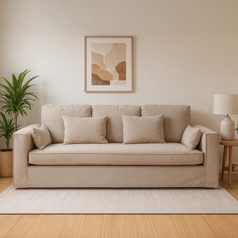 Three-seat grey fabric sofa with plush cushions, set against a light-colored wall. A framed artwork hangs above the sofa, and a potted plant is positioned to the left. A soft area rug is placed underneath the sofa, enhancing the cozy atmosphere of the room