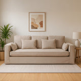 Three-seat grey fabric sofa with plush cushions, set against a light-colored wall. A framed artwork hangs above the sofa, and a potted plant is positioned to the left. A soft area rug is placed underneath the sofa, enhancing the cozy atmosphere of the room
