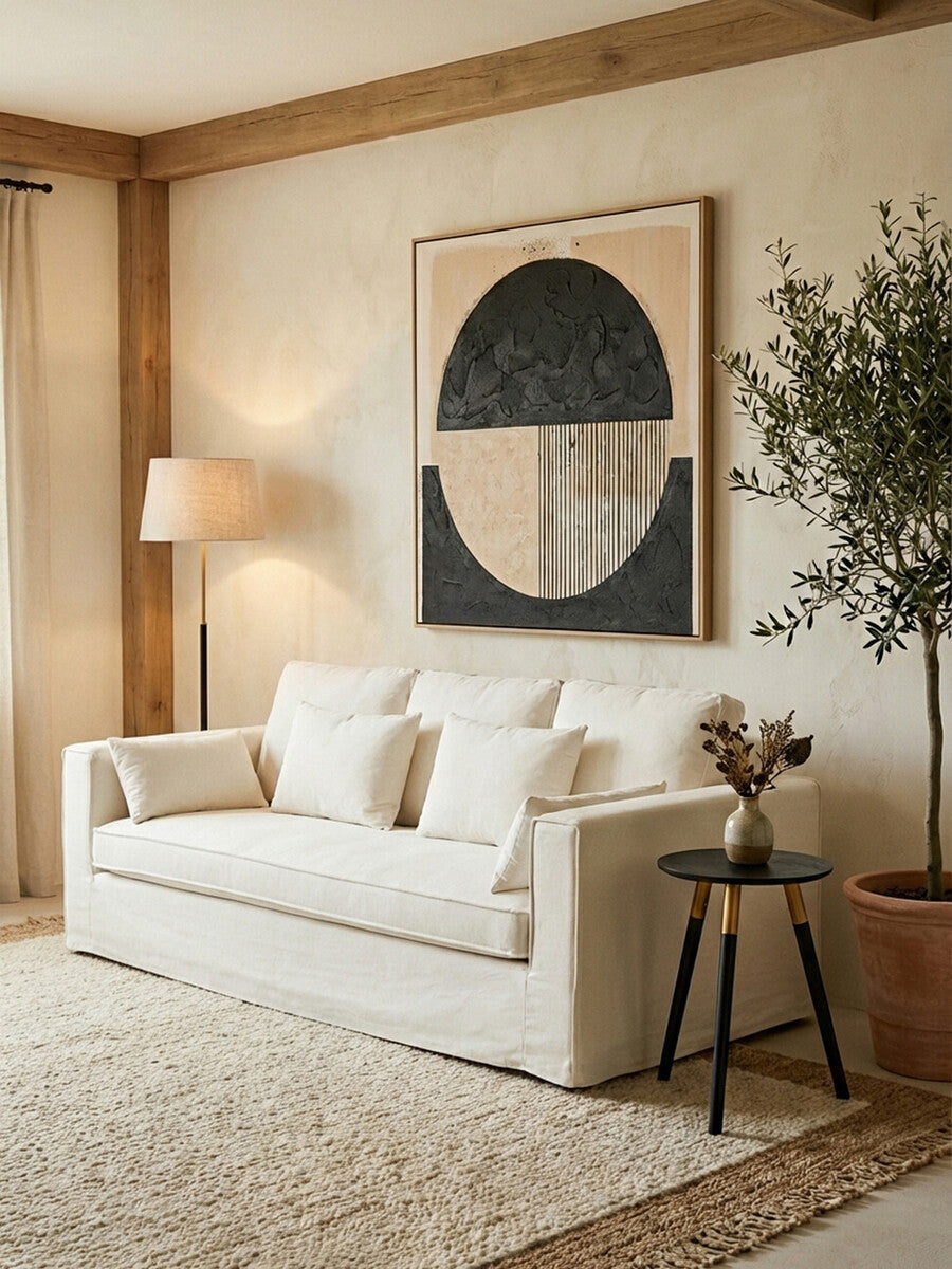 A cream-colored three-seat sofa with plush cushions is positioned against a light beige wall. A large abstract artwork featuring geometric shapes hangs above the sofa. To the right, a tall indoor plant adds greenery, and a small black side table with a decorative vase sits beside the sofa. A textured area rug covers the floor, enhancing the cozy atmosphere of the room