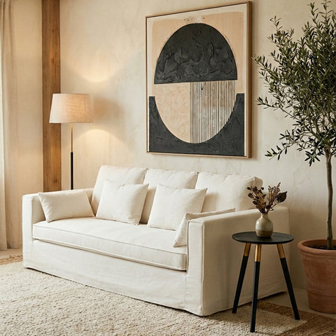 Cream-colored three-seat sofa with plush cushions, positioned in a cozy living room setting. A modern abstract artwork hangs on the wall behind the sofa, and a stylish floor lamp stands nearby. A small round side table with a decorative vase and a plant adds to the inviting atmosphere. The floor is covered with a light-colored rug, enhancing the warm ambiance of the space