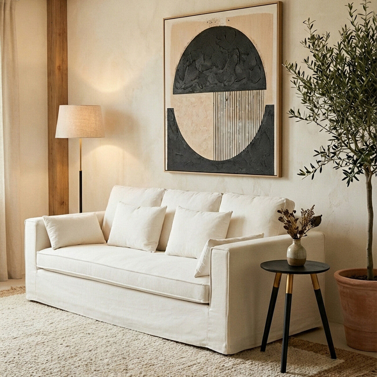 Cream-colored three-seat sofa with plush cushions, positioned in a cozy living room setting. A modern abstract artwork hangs on the wall behind the sofa, and a stylish floor lamp stands nearby. A small round side table with a decorative vase and a plant adds to the inviting atmosphere. The floor is covered with a light-colored rug, enhancing the warm ambiance of the space