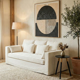 Cream-colored three-seat sofa with plush cushions, positioned in a cozy living room setting. A modern abstract artwork hangs on the wall behind the sofa, and a stylish floor lamp stands nearby. A small round side table with a decorative vase and a plant adds to the inviting atmosphere. The floor is covered with a light-colored rug, enhancing the warm ambiance of the space
