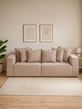 Three-seat grey sofa with plush cushions, positioned against a light-colored wall. Two framed artworks hang above the sofa, and a small side table with a lamp is visible to the right. A green plant is placed beside the sofa, and a light area rug covers the wooden floor