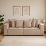 Three-seat grey sofa with plush cushions, set against a light-colored wall. Two framed artworks hang above the sofa, and a potted plant is visible on the left. A light-colored rug is placed underneath, complementing the overall neutral color scheme of the room