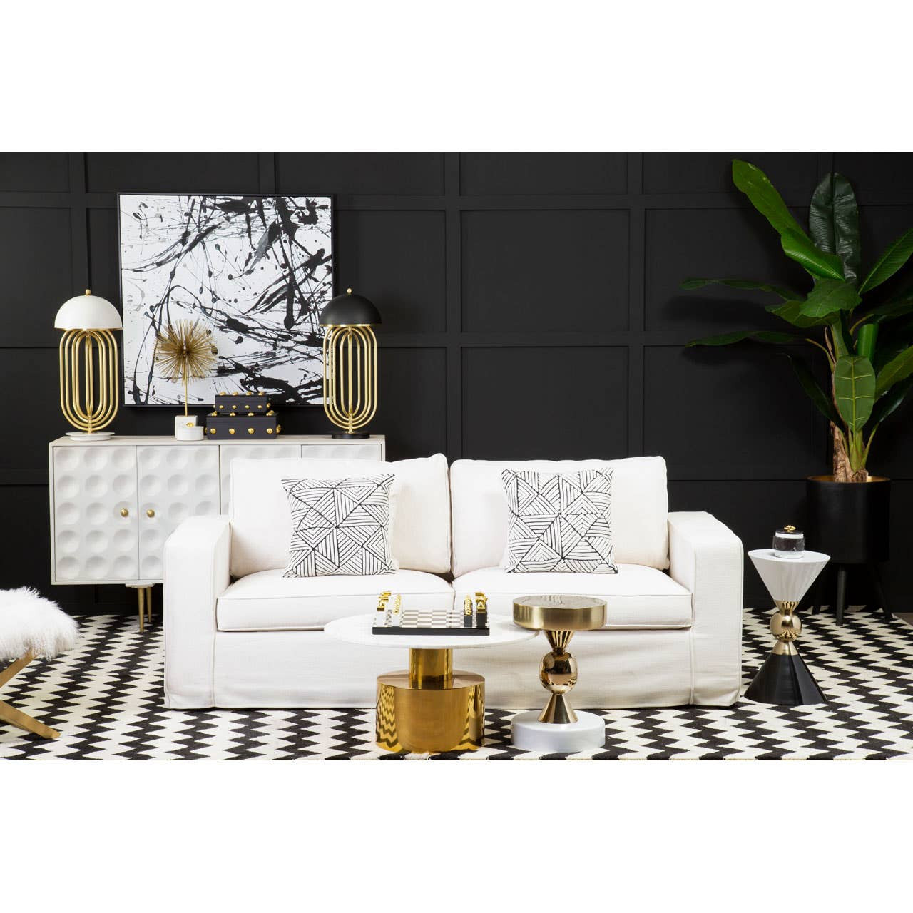 A cream fabric sofa with three seats, featuring decorative cushions, is positioned in a stylish living room setting. The backdrop includes a black wall adorned with abstract artwork, while a striped rug lies beneath the sofa. A modern side table and a decorative plant are also visible, adding to the contemporary decor