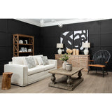 A cream fabric sofa with three seats is positioned in a modern living room setting. The sofa features decorative cushions and is complemented by a wooden coffee table with a decorative plant on top. Behind the sofa, there is a black accent wall with abstract art, and to the side, a wooden bookshelf displays various decor items. Two stylish lamps are placed on either side of the bookshelf, and a unique chair is