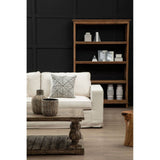 Cream fabric sofa with three seats, featuring a decorative gray patterned throw pillow. In the background, a wooden bookshelf displays various decorative items, including vases and books, against a dark wall. A wooden coffee table is positioned in front of the sofa, adding to the cozy living room setting