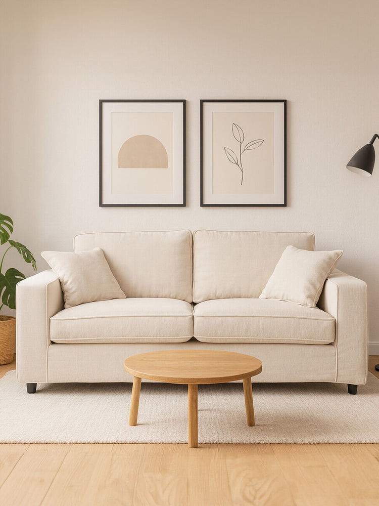 A cream fabric two-seat sofa with plush cushions, positioned against a light-colored wall. Above the sofa, there are two framed art prints featuring simple line drawings. A small round wooden coffee table sits in front of the sofa, and a potted plant is visible to the left, adding a touch of greenery to the scene. The flooring is light wood, enhancing the cozy atmosphere
