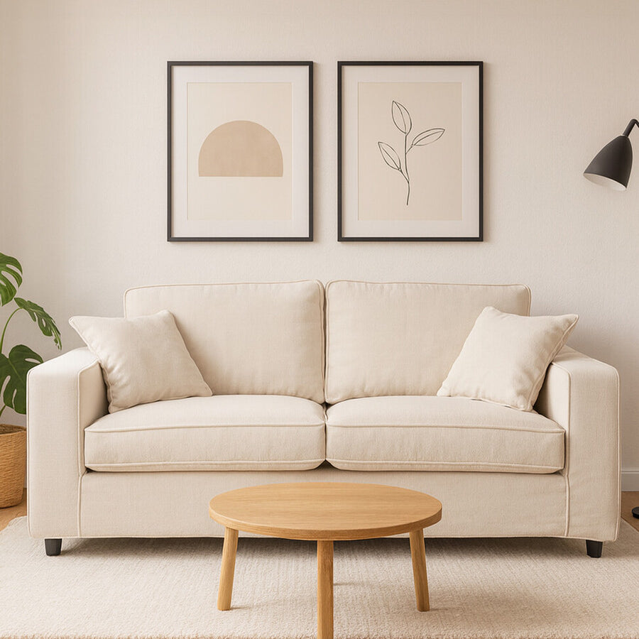 A cream fabric two-seat sofa with plush cushions, positioned against a light-colored wall. Above the sofa, there are two framed art prints featuring simple line drawings. A small round wooden coffee table sits in front of the sofa, and a potted plant is visible to the left, adding a touch of greenery to the scene. The flooring is light wood, enhancing the cozy atmosphere
