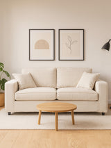 A cream fabric two-seat sofa with plush cushions, positioned against a light-colored wall. Above the sofa, there are two framed art prints featuring simple line drawings. A small round wooden coffee table sits in front of the sofa, and a potted plant is visible to the left, adding a touch of greenery to the scene. The flooring is light wood, enhancing the cozy atmosphere