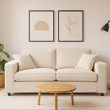 Cream fabric two-seat sofa with plush cushions, positioned against a light-colored wall. Above the sofa are two framed art prints featuring simple geometric and botanical designs. A small round wooden coffee table is placed in front of the sofa, and a potted plant is visible to the left. A modern floor lamp stands to the right, adding a warm ambiance to the scene